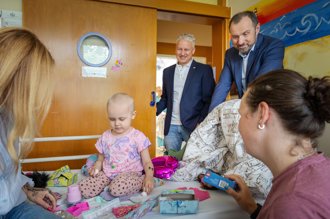 Dítě na posteli obklopené hračkami v pozadí Erich Stavař a Jakub Vaněk